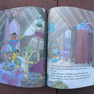May include: A colorful illustration from a children's book depicts a cluttered attic scene. Two Sesame Street characters, Bert and Ernie, are exploring the attic. The image includes a gramophone, lamps, a framed portrait, and a wardrobe.