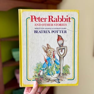 May include: A vintage hardcover book titled "Peter Rabbit and Other Stories" by Beatrix Potter. The cover features an illustration of Peter Rabbit in a blue jacket eating a carrot, with a bird perched on a shovel. The book has a yellow border.