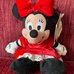 May include: A red plush Minnie Mouse wearing a red velvet dress with white lace trim and a red bow. The plush toy has a Disney Store tag attached.