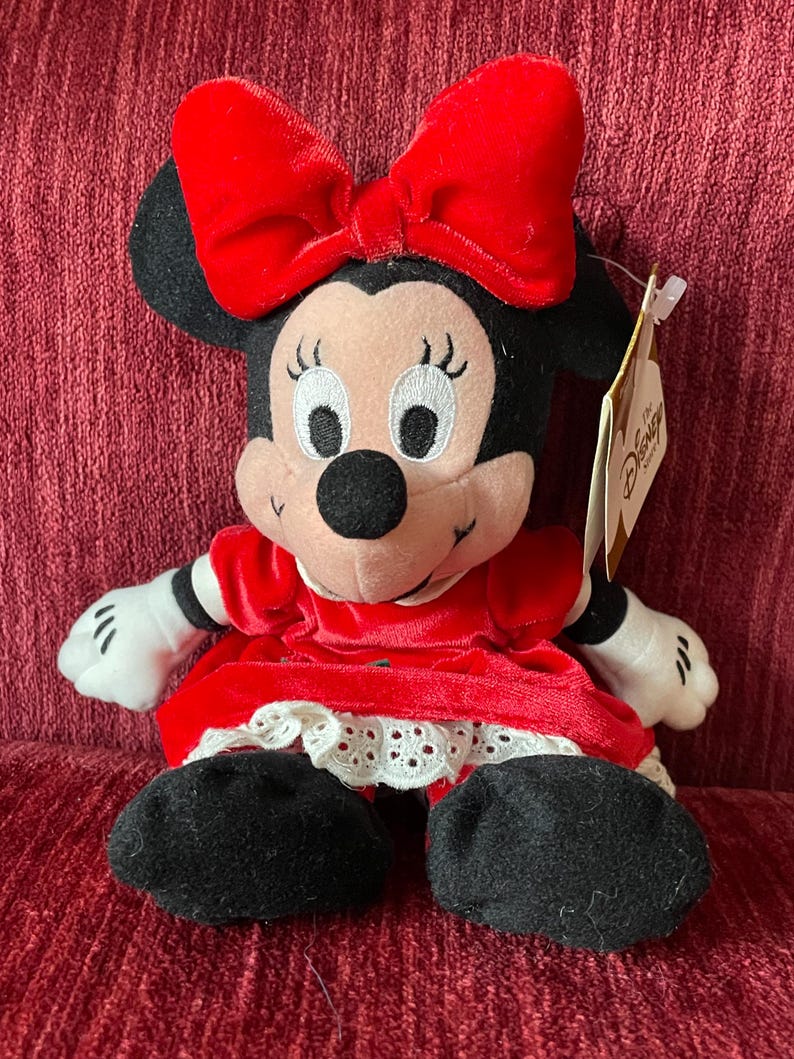 May include: A red plush Minnie Mouse wearing a red velvet dress with white lace trim and a red bow. The plush toy has a Disney Store tag attached.