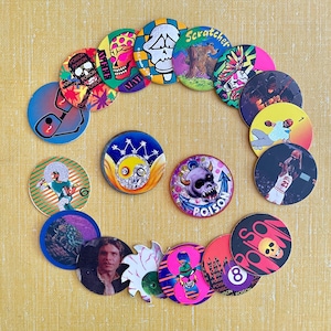 May include: A collection of circular cardboard pogs featuring various graphic designs including skulls, sports figures, a portrait of a man, a yin-yang symbol, and the word Poison. The colorful discs are arranged in a spiral pattern on a yellow surface.