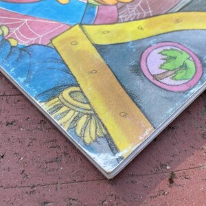 May include: A corner of a colorful children's book illustration. The image features a blue and yellow section with a circular emblem of a palm tree. The book's edge is visible, showing wear and tear.