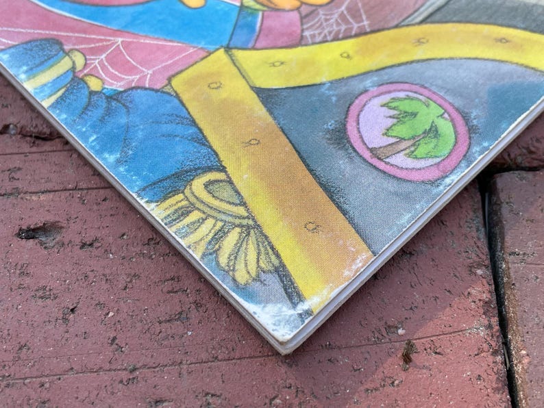 May include: A corner of a colorful children's book illustration. The image features a blue and yellow section with a circular emblem of a palm tree. The book's edge is visible, showing wear and tear.