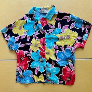 May include: Short-sleeved button-up shirt with a vibrant floral pattern. The shirt features large flowers in shades of red, yellow, and blue against a black background. The collar and sleeves are trimmed in a light blue color. The shirt is made of a lightweight fabric.