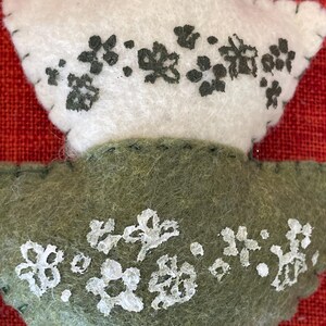 K&ouml;nnte beinhalten: Zwei Filzornamente in Form von Bl&auml;ttern. Das obere Ornament ist wei&szlig; mit schwarzen gestickten Blumen. Das untere Ornament ist gr&uuml;n mit wei&szlig;en gestickten Blumen.