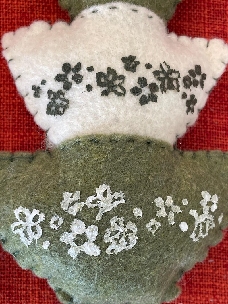 K&ouml;nnte beinhalten: Zwei Filzornamente in Form von Bl&auml;ttern. Das obere Ornament ist wei&szlig; mit schwarzen gestickten Blumen. Das untere Ornament ist gr&uuml;n mit wei&szlig;en gestickten Blumen.