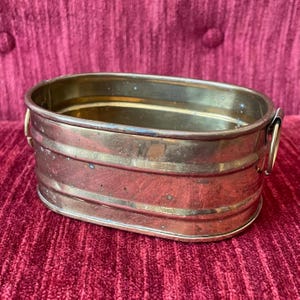 May include: An antique brass planter with an oval shape and two small ring handles. The planter has a warm, aged patina and horizontal bands around the sides. The interior is a lighter brass color.