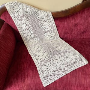 May include: Cream-colored, open-weave lace table runner with a floral pattern. The runner is draped over a red sofa, showcasing the intricate design and texture of the lace. A decorative piece for home decor.