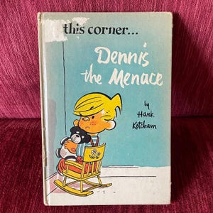 May include: Vintage hardcover book titled "Dennis the Menace" by Hank Ketcham. The cover features an illustration of Dennis the Menace holding a teddy bear while sitting in a yellow rocking chair. The background is light blue with the words "this corner..." at the top.