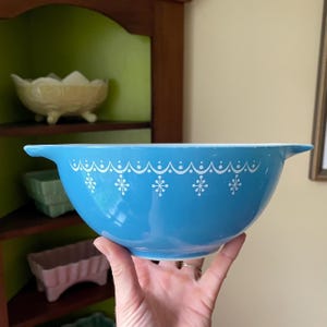 May include: A vintage, light blue mixing bowl with a white floral and dotted pattern around the rim. The bowl has a handle on each side. This retro kitchenware is perfect for baking and serving.
