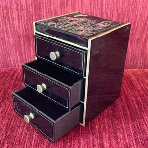 May include: A black tiered storage chest with three drawers, each featuring a small round brass-colored knob. The top surface displays a detailed floral illustration with butterflies. The unit has thin gold-toned trim along the edges.