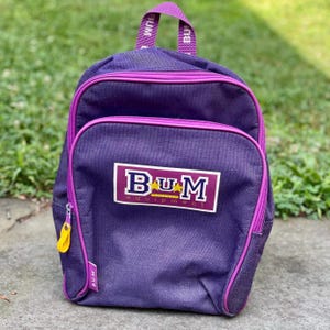 May include: Purple backpack with a zippered front pocket and a top handle. The backpack has a rectangular patch with the text "BUM Equipment" in white and gold. The zipper pull is yellow, and the trim is a bright pink.