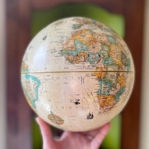 May include: A vintage globe with a cream-colored surface, displaying continents and countries in various colors. The globe features detailed cartography, including ocean names and illustrations of ships. The globe is held in a person's hand.