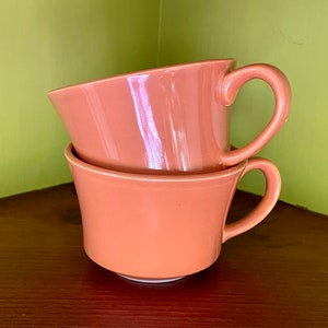 May include: Two stacked peach-colored ceramic teacups with curved handles. The cups have a flared shape and sit on a dark wooden surface against a green wall. The cups are empty and appear to be clean.