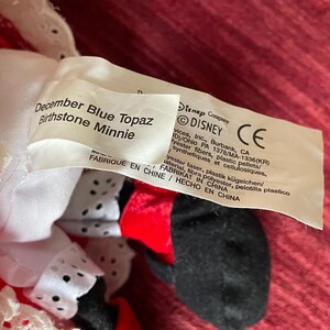 May include: A plush Minnie Mouse toy wearing a red dress with white lace trim and black polka dots. The tag reads "December Blue Topaz Birthstone Minnie" and includes information about the Disney Company.