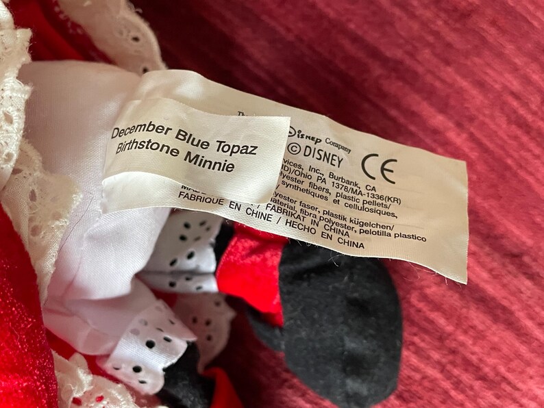 May include: A plush Minnie Mouse toy wearing a red dress with white lace trim and black polka dots. The tag reads "December Blue Topaz Birthstone Minnie" and includes information about the Disney Company.