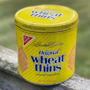 May include: A cylindrical yellow metal tin for Wheat Thins snack crackers. The label features the Nabisco logo, the text Original Wheat Thins, Limited Edition, and whole wheat goodness. The container displays a net weight of 13 oz.