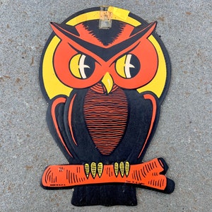 May include: A vintage Halloween decoration featuring a black and orange owl with large, yellow eyes. The owl is perched on a branch and has a yellow moon behind it.