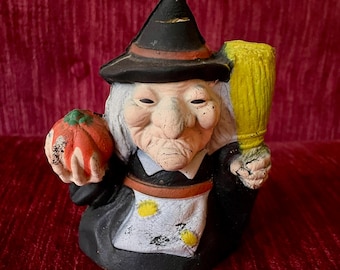 May include: A vintage Halloween witch figurine with a black hat, white hair, and a broom. She is holding a red tomato in her left hand and has a grumpy expression on her face.