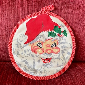 May include: A round Christmas pot holder featuring a Santa Claus face. The pot holder has a red border with a white and red checkered trim. Santa's hat is red, and his face is a light peach color with a white beard. Holly and berries are on the hat.
