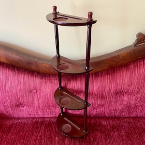 May include: A dark brown wooden wall shelf featuring four semi-circular tiers. Each tier has a smooth finish, a recessed circular indentation, and a narrow rectangular slot. The unit is supported by two vertical wooden poles with small finials.