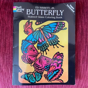 May include: A Dover Coloring Book titled Butterfly Stained Glass Coloring Book by Ed Sibbett, Jr. The cover features a black background with a central yellow and orange panel displaying four colorful butterflies in shades of blue, pink, and green.