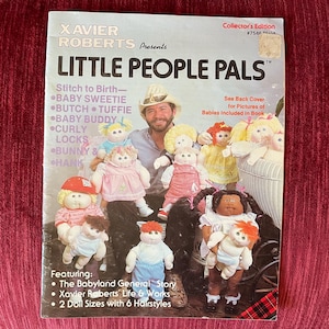 May include: A vintage "Little People Pals" book cover featuring a man surrounded by several dolls. The cover has text that reads "Xavier Roberts Presents" and "Collector's Edition." The dolls have various hairstyles and outfits.