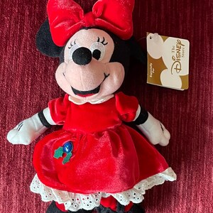May include: Red plush Minnie Mouse wearing a red velvet dress with white lace trim and a red bow. She has a green holly sprig on her dress with a blue button. The tag reads "The Disney Store" and "Especially For".