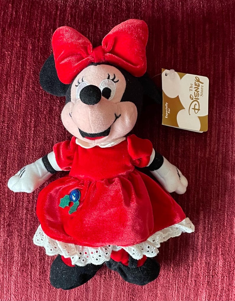 May include: Red plush Minnie Mouse wearing a red velvet dress with white lace trim and a red bow. She has a green holly sprig on her dress with a blue button. The tag reads "The Disney Store" and "Especially For".