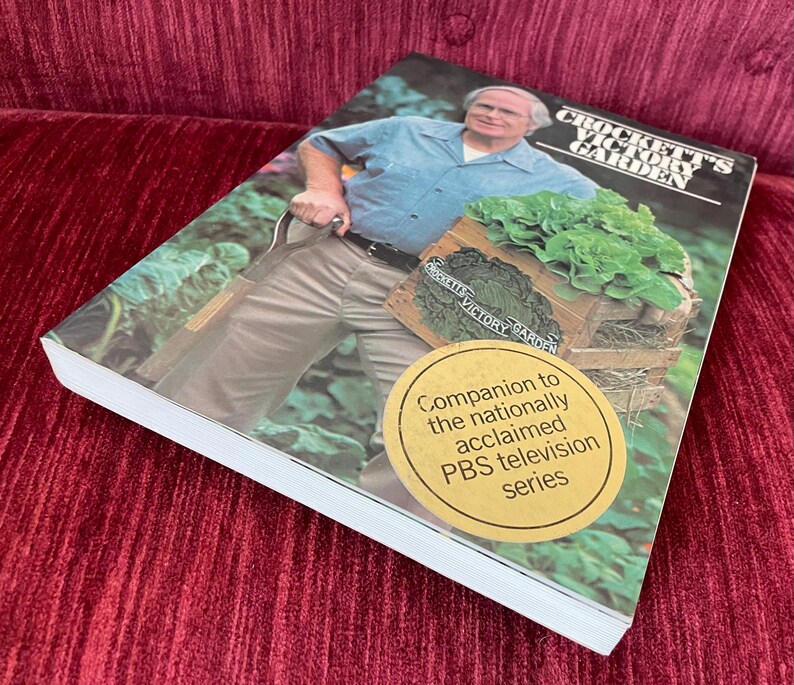 May include: A softcover book titled Crockett's Victory Garden featuring a photograph of a person in a blue shirt and tan pants holding a wooden crate of lettuce. A round gold sticker on the cover reads Companion to the nationally acclaimed PBS television series.