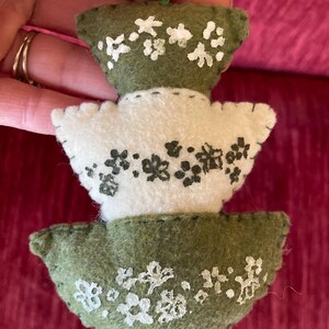 K&ouml;nnte beinhalten: Ein Filz-Weihnachtsbaum-Anh&auml;nger mit drei Ebenen. Jede Ebene hat eine andere Farbe: gr&uuml;n, wei&szlig; und gr&uuml;n. Der Anh&auml;nger ist mit wei&szlig;en und schwarzen Blumendesigns verziert.
