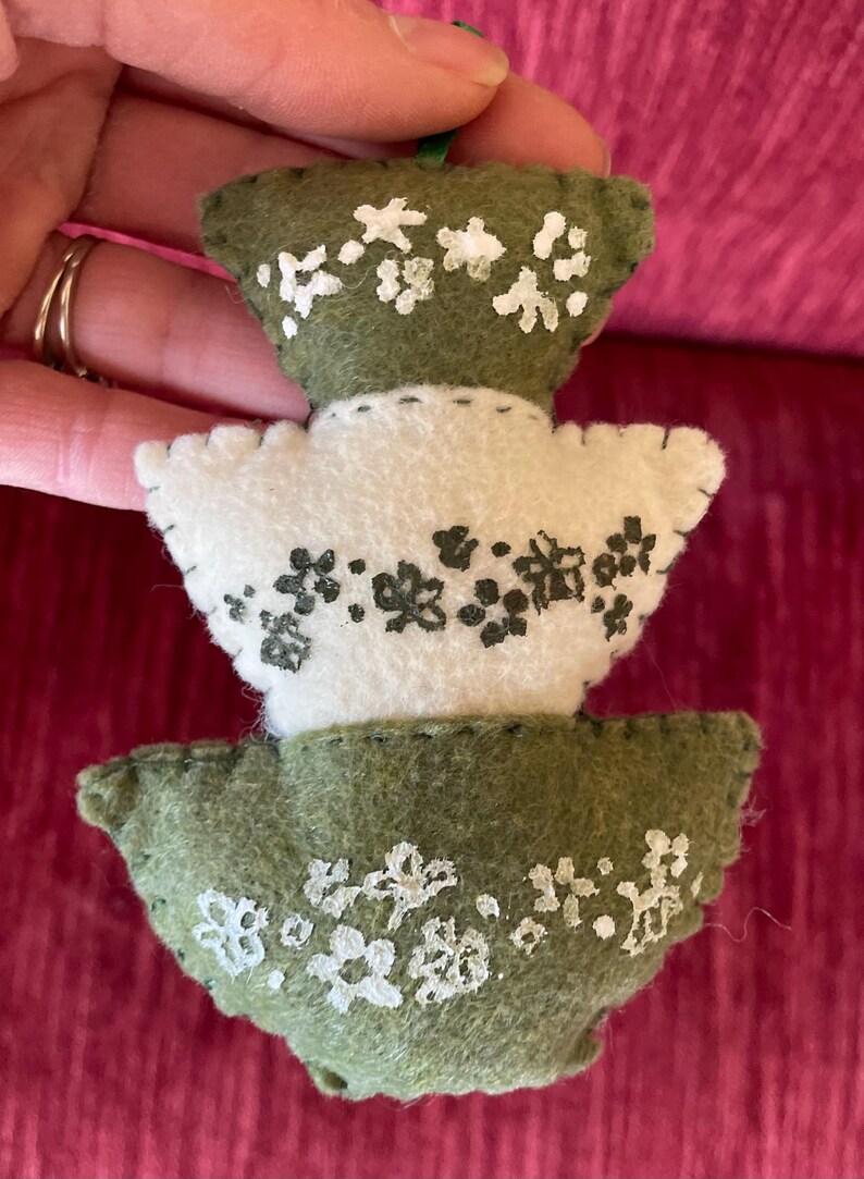 K&ouml;nnte beinhalten: Ein Filz-Weihnachtsbaum-Anh&auml;nger mit drei Ebenen. Jede Ebene hat eine andere Farbe: gr&uuml;n, wei&szlig; und gr&uuml;n. Der Anh&auml;nger ist mit wei&szlig;en und schwarzen Blumendesigns verziert.