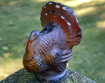 May include: A ceramic turkey figurine with a brown and black body and a red, white, and brown tail feathers. The turkey is facing to the right and has its head turned back to look over its shoulder.