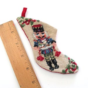 May include: A small, hand-stitched Christmas stocking featuring a nutcracker soldier design. The stocking is beige with red, green, blue, and black accents. A red loop is attached for hanging. A wooden ruler is next to the stocking.