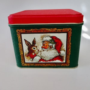 May include: A vintage Christmas tin with a red lid and green body. The tin features a framed illustration of Santa Claus and a deer. The tin is likely used for storing holiday treats or small gifts.