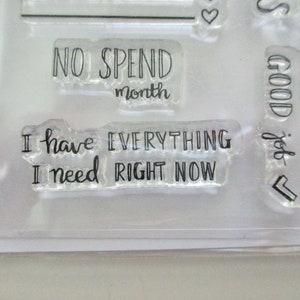 May include: Clear acrylic stamps with black text. Phrases include "NO SPEND month" and "I have EVERYTHING I need RIGHT NOW." Other stamps include the words "GOOD job" and a heart symbol.