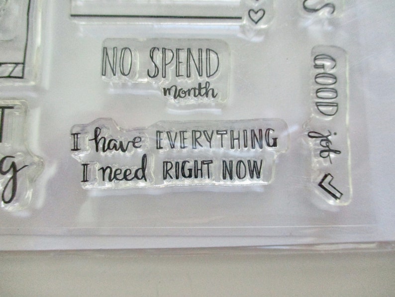 May include: Clear acrylic stamps with black text. Phrases include "NO SPEND month" and "I have EVERYTHING I need RIGHT NOW." Other stamps include the words "GOOD job" and a heart symbol.