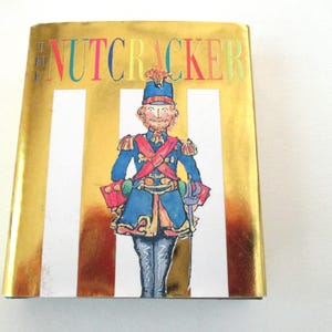 May include: A hardcover book titled The Nutcracker with a shiny gold metallic cover featuring vertical white stripes. The center displays a colorful illustration of a soldier in a blue uniform with red accents, a tall hat, and a sword at their side.