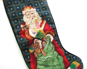 May include: A needlepoint Christmas stocking featuring a dark blue plaid background with a central figure of Santa Claus wearing a red coat and white beard, holding a green sack filled with toys including a brown teddy bear and a doll.