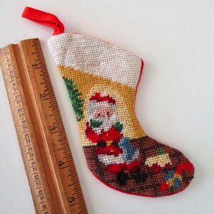 May include: Miniature Christmas stocking with a Santa Claus design. The stocking is made of needlepoint with a red trim and a red ribbon loop. A wooden ruler is next to the stocking.