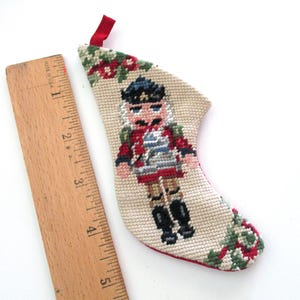May include: Miniature Christmas stocking with a cross-stitch design of a nutcracker soldier. The stocking is beige with red, blue, black, and green accents. A wooden ruler is next to the stocking.