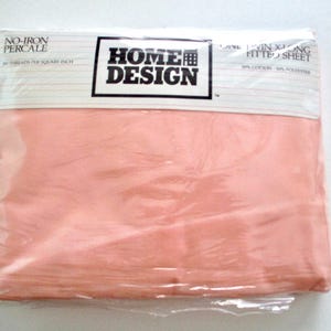May include: A package of Home Design twin-size fitted sheet in a light peach color. The packaging is clear plastic, and the sheet is visible inside. The top of the package has the words "No-Iron Percale" and "Home Design" printed on it.