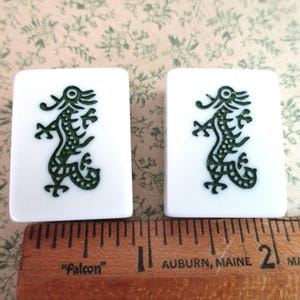 May include: Two white rectangular Mahjong tiles, each featuring a dark green dragon design. The tiles are next to a wooden ruler with inch markings, showing the tiles' size.