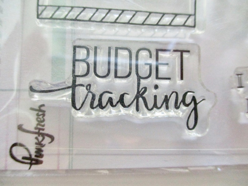 May include: Clear acrylic stamp with the words "BUDGET tracking" in black, handwritten-style font. The stamp is part of a set, with a partial view of another stamp at the top. The brand name "Pinkfresh" is visible in the lower left corner.
