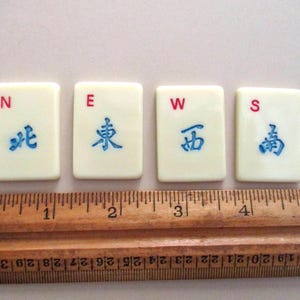 May include: Four off-white Mahjong tiles with red and blue markings. The tiles are labeled N, E, W, and S, with corresponding Chinese characters. A wooden ruler is placed below the tiles, measuring in inches.