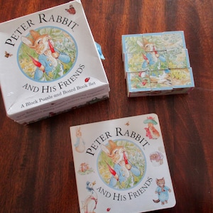 May include: A Peter Rabbit and Friends block puzzle and board book set. The set includes a white box with a blue ribbon, a small book, and a cube puzzle. The illustrations feature Peter Rabbit and other characters.