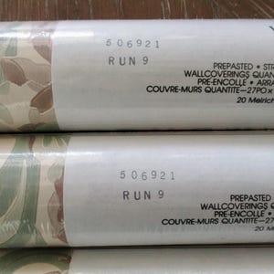May include: Close-up of wallpaper rolls with a camouflage-like pattern in shades of green, brown, and beige. The rolls have white labels with the text "506921 RUN 9" and other details. The wallpaper is pre-pasted and strippable.