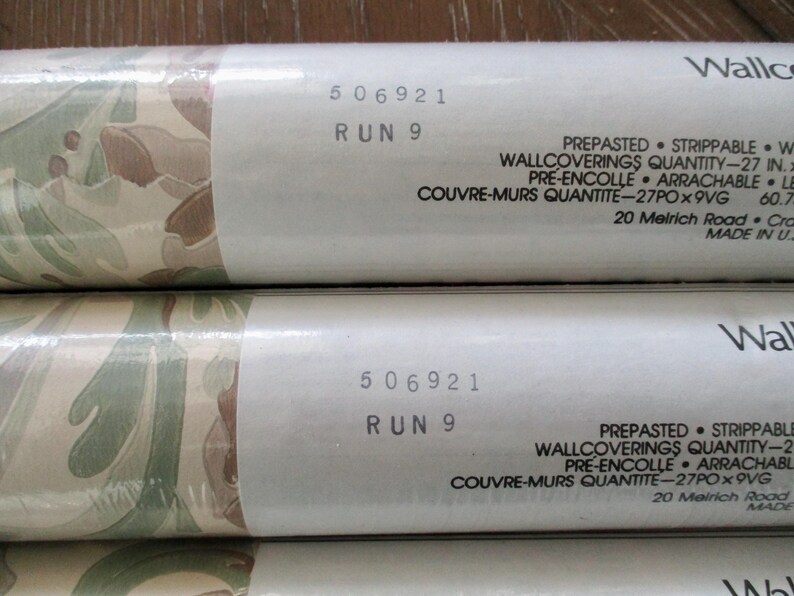 May include: Close-up of wallpaper rolls with a camouflage-like pattern in shades of green, brown, and beige. The rolls have white labels with the text "506921 RUN 9" and other details. The wallpaper is pre-pasted and strippable.