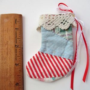 May include: A miniature Christmas stocking, approximately 3 inches long, crafted from various patterned fabrics. The stocking features red and white stripes, blue gingham, and green plaid. It is trimmed with a cream-colored lace and has red and white ribbons for hanging.