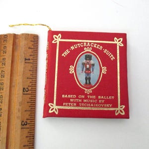May include: A small red book titled The Nutcracker Suite based on the ballet with music by Peter Tschaikovsky, featuring a gold-bordered illustration of a nutcracker. A wooden ruler shows the book is approximately 3 inches in height.